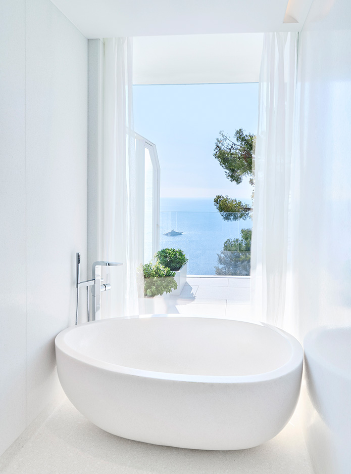 A spacious round bathtub next to a glass wall with a coastline view.