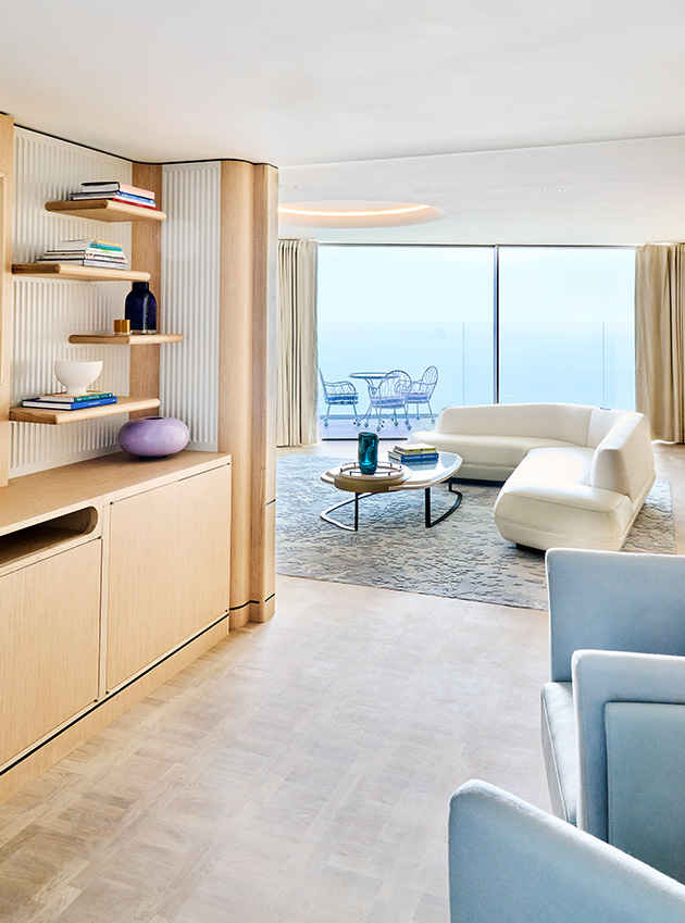 Large curved white and blue sofas surround a sleek  glass coffee table and a wall of art and books in the Grand Sea View Suite sitting area