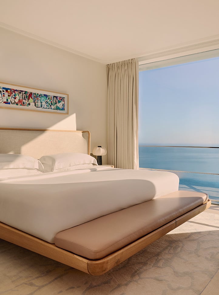 Bright, modern bedroom with a large bed dressed in white linens, a padded leather bench at the foot, and a floor-to-ceiling window offering uninterrupted views of the calm blue sea, with soft natural light streaming across the room.