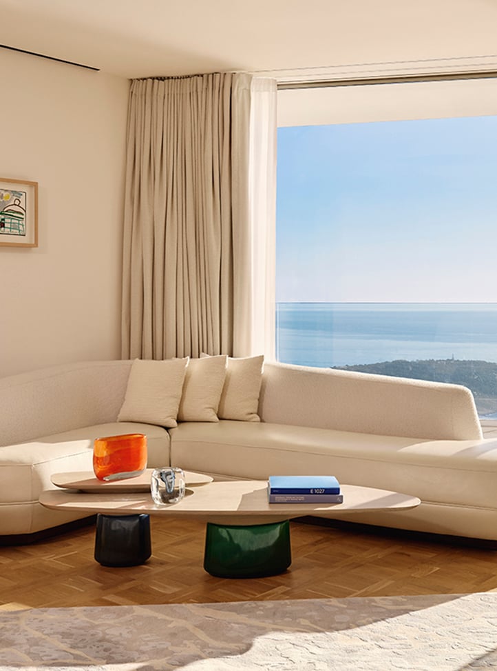 Elegant living area with a curved cream sofa and neutral cushions, two sculptural coffee tables with colourful bases, and a large window offering sweeping sea views, framed by floor-length curtains and parquet flooring.