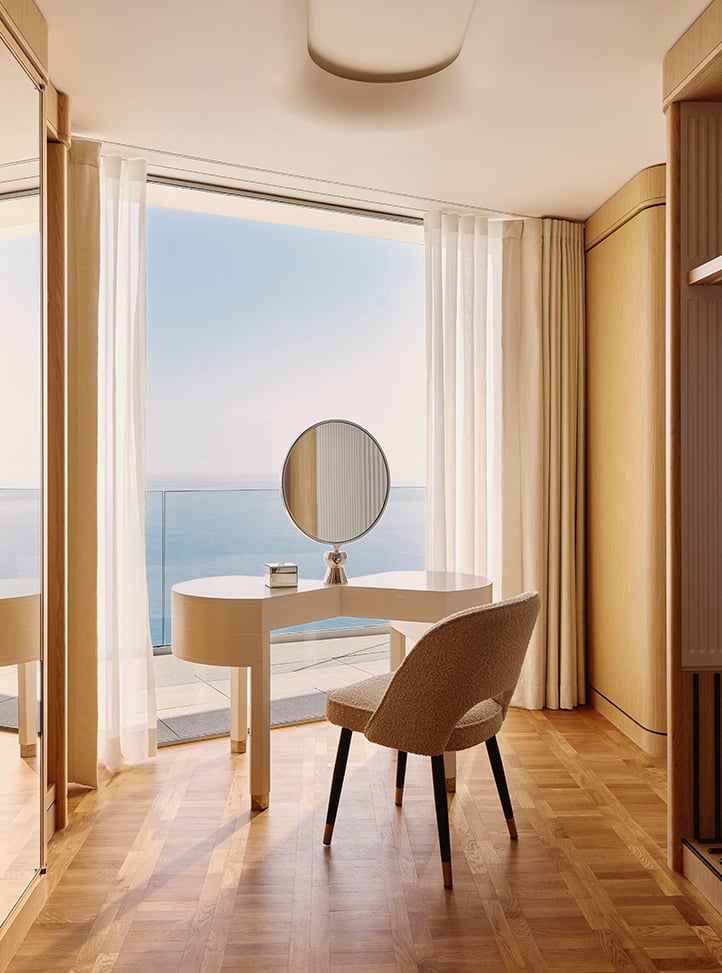 Elegant dressing area with a curved white vanity table, round mirror, and upholstered chair, positioned in front of floor-to-ceiling windows overlooking a calm sea, with sheer curtains and warm wood flooring.