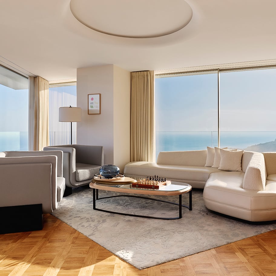Sophisticated living room with a curved cream sofa, matching armchair, and a modern oval coffee table set on a patterned rug, featuring a chess set and glass bowl, with floor-to-ceiling windows offering panoramic sea and coastal views.