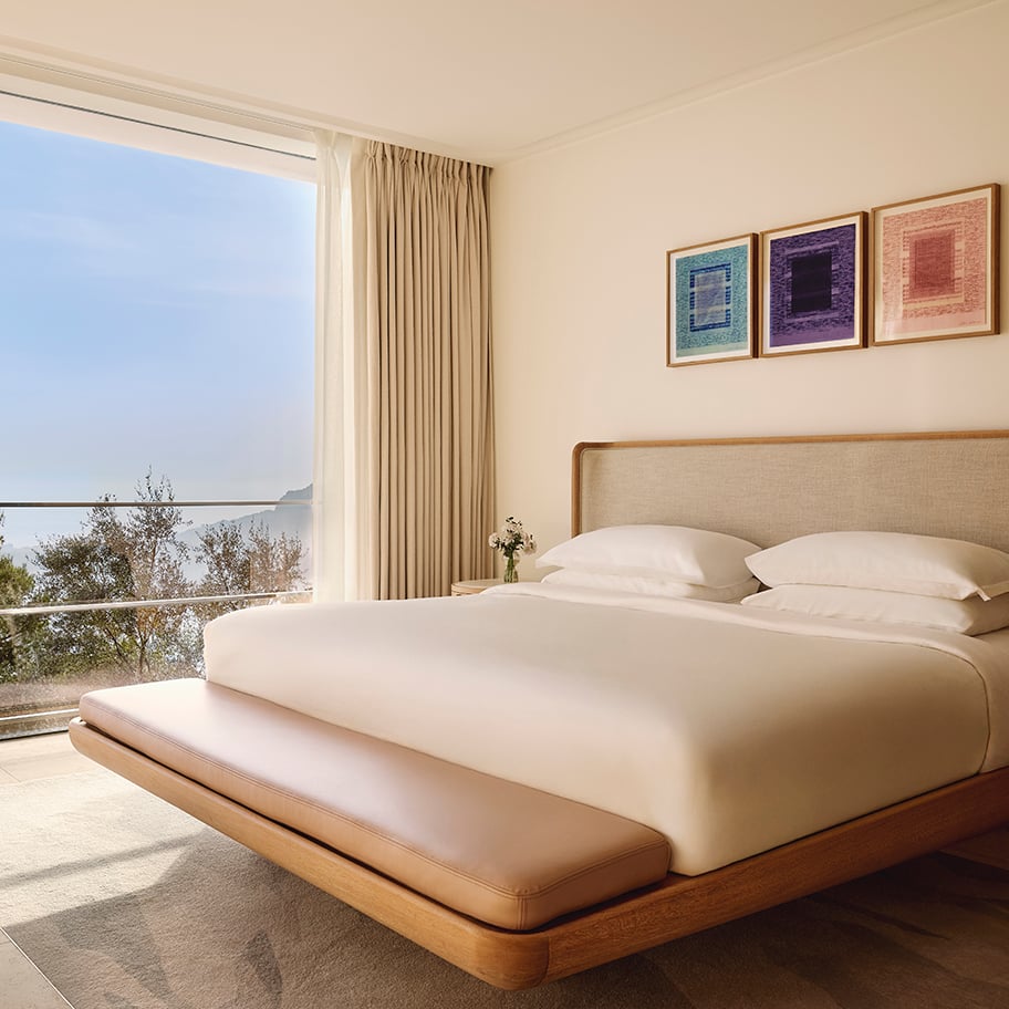 Serene bedroom with a large bed featuring white linens and a wooden frame, a cushioned bench at the foot, and a floor-to-ceiling window offering views of the sea and trees, with three colourful abstract artworks above the headboard.