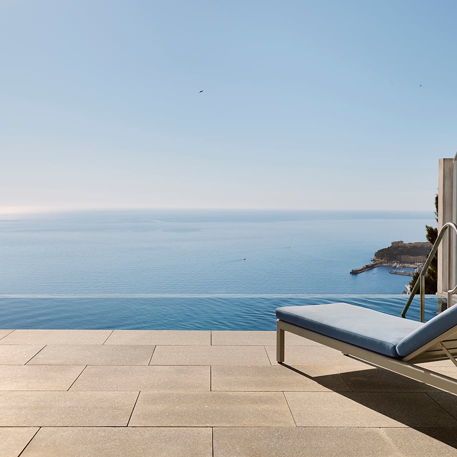 Tranquil terrace view with a single cushioned sun lounger beside an infinity pool, overlooking the calm blue sea and a distant coastline under a clear sky.