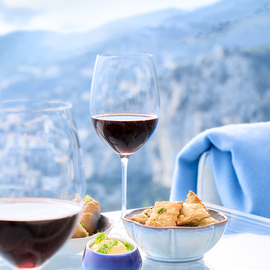 Deux verres de vin rouge posés sur une table avec des biscuits apéritif, et la vue sur la Côte d'Azur an arrière-plan.