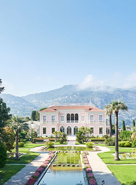 An elongated fountain above the grass with a villa in the background that has a mountain behind it.