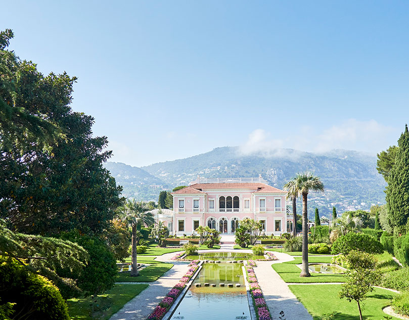 An elongated fountain above the grass with a villa in the background that has a mountain behind it.