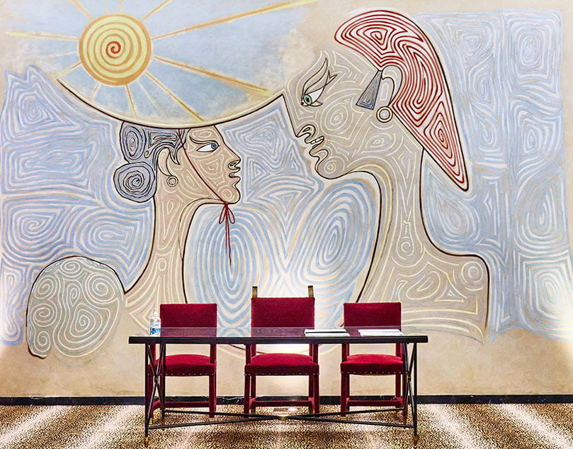A table with three red chairs behind it, and on the wall an artistic painting.