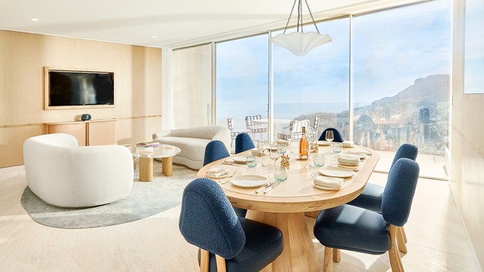 A living room with a dining table and chairs around, and a sitting area with a sofa and armchairs. In the background is a floor-to-ceiling window with a view on the Riviera