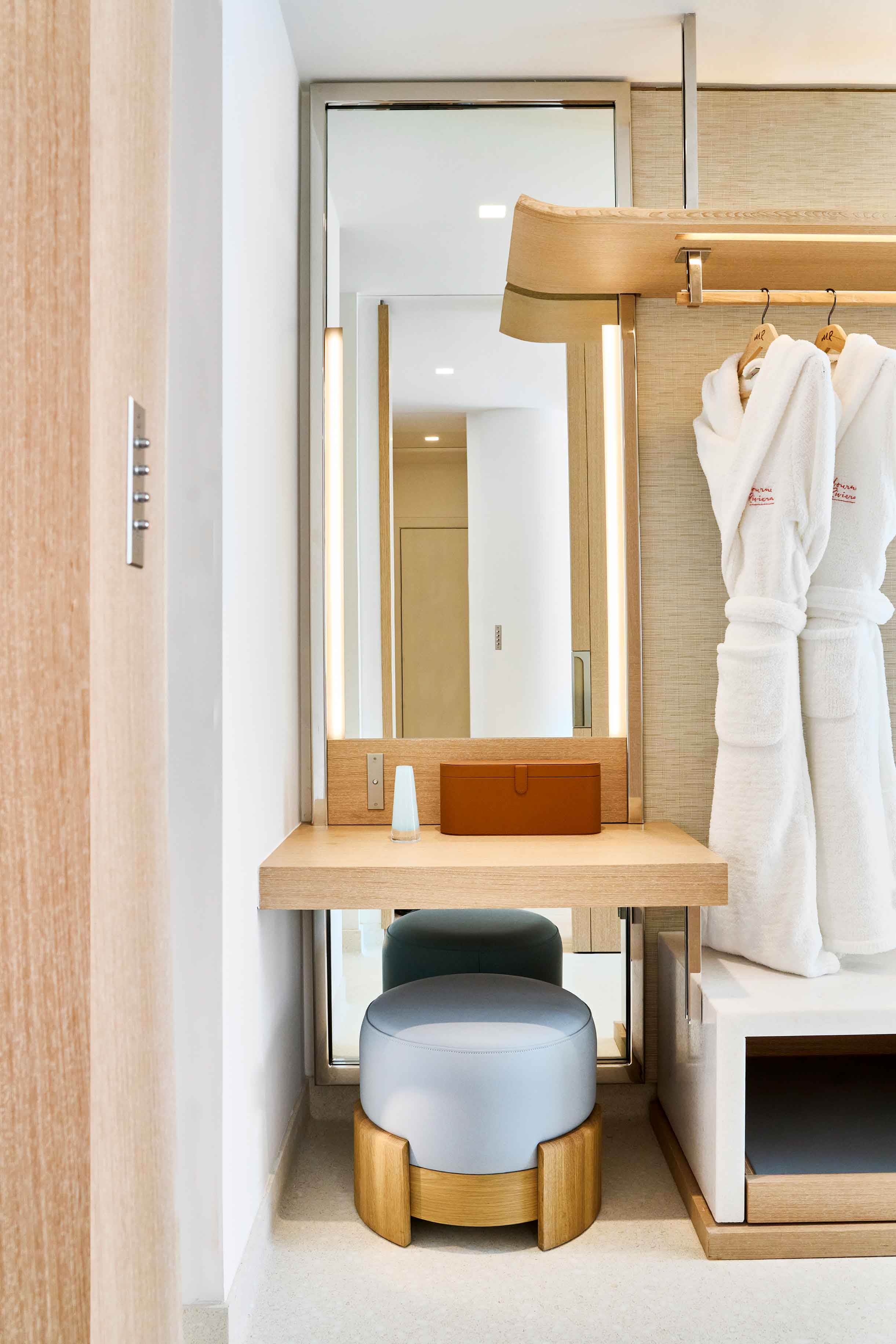 A wooden dressing table with an armchair and a long mirror next to two white bathrobes.