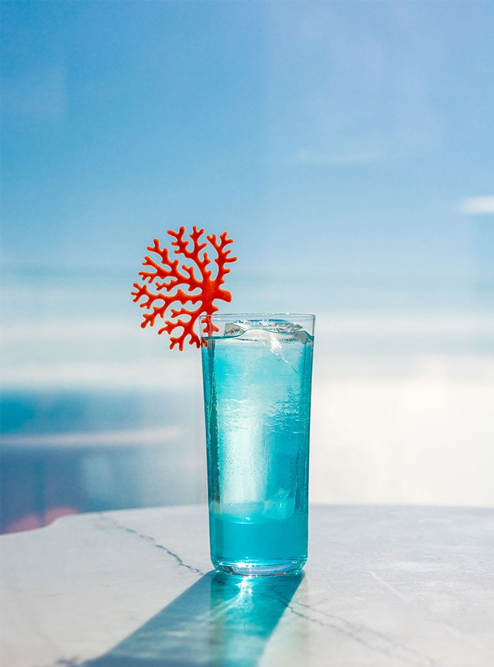 Tall blue cocktail with coral garnish in front of sea