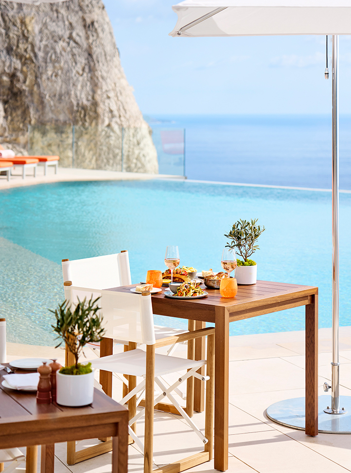 La Piscine by Jean-Georges au Maybourne Riviera - table en bois avec deux chaises blanches de part et d'autre et des plats servis sur la table. Derrière la table se trouve la piscine avec la vue sur la mer à l'infini.