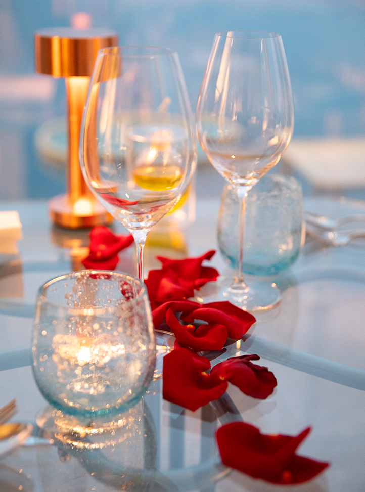 Red rose petals on a glass table with wine and water glasses.