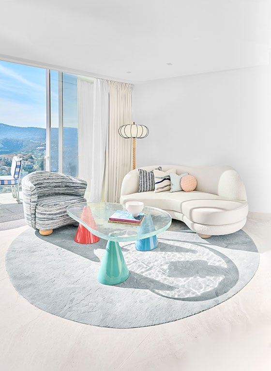 The Sea View Duplex Suite features a curved cream fabric sofa behind a glass coffee table with blue and red table legs, the room also has a balcony with a blue and white striped chair