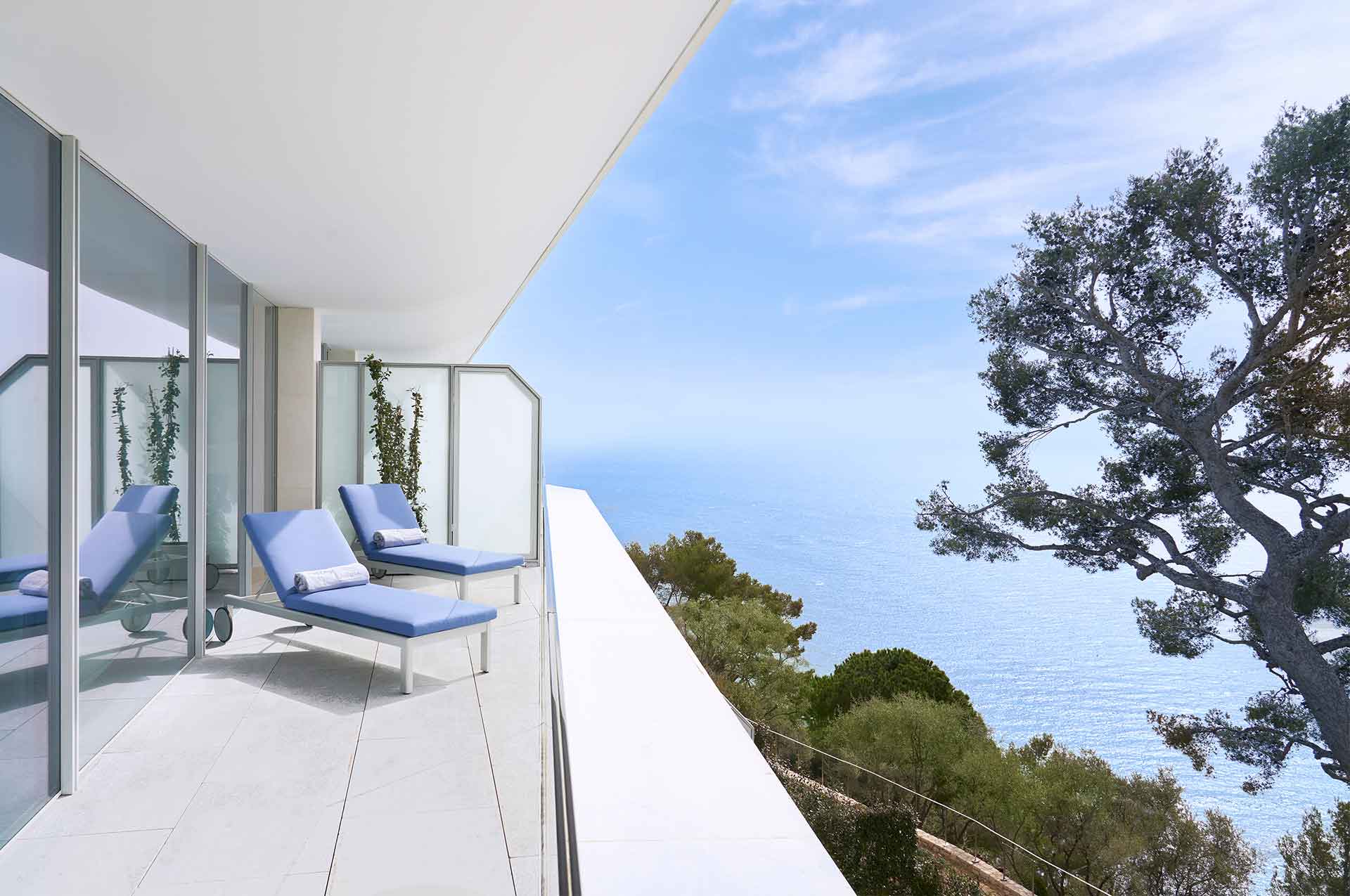 The Sea View Suite balcony overlooks the blue waters and lush nature surrounding the property. Two blue deckchairs rest on the balcony looking over a large curved tree in the garden
