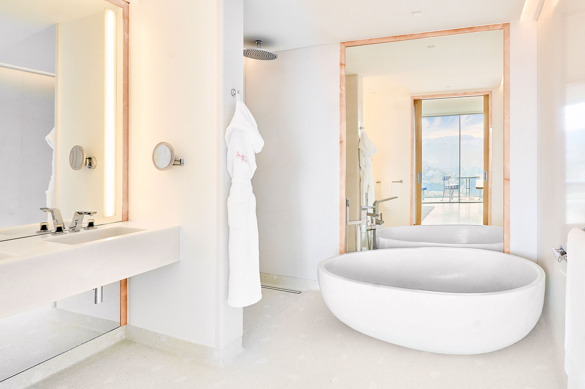 The wide curved bath sits in the centre of the modern Sea View Suite bathrooms, with branded Maybourne Riviera dressing robes hanging from the hooks beside the shower