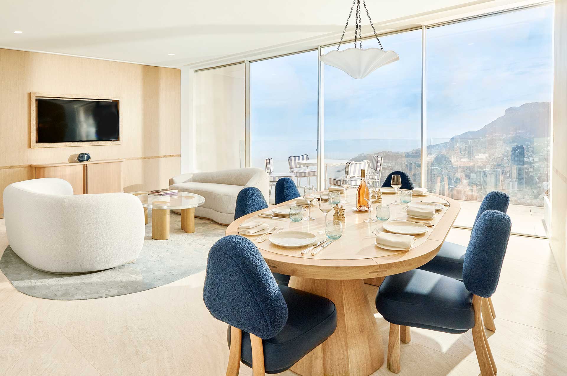 The Sea View Suite lounge features curved white plush sofas, a long wooden dining table surrounded by modern blue chairs all overlooking panoramic views of Monaco from the balcony