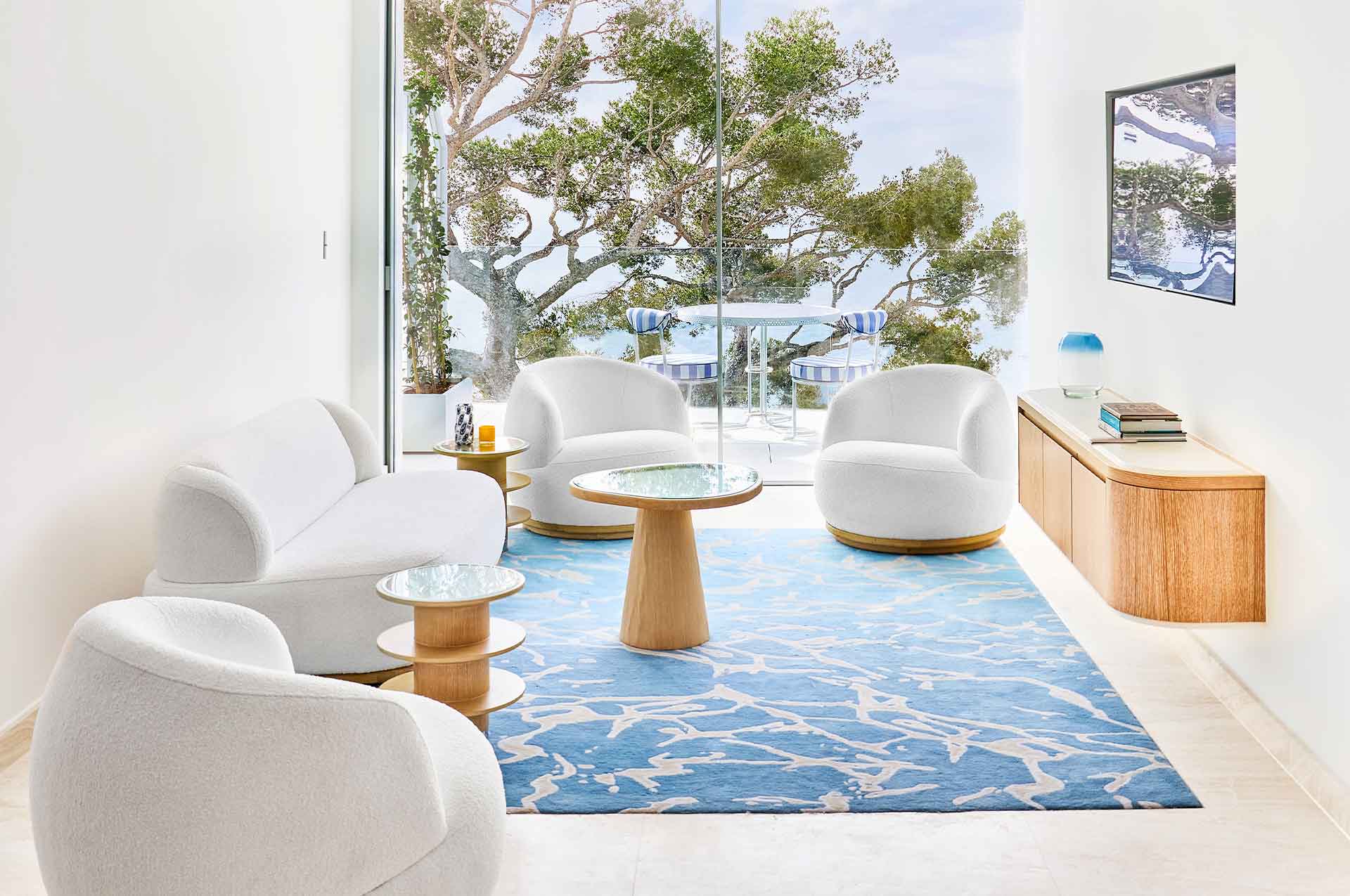 The Sea View Suite lounge features 4 grey rounded sofas and armchairs surrounding curved wooden tables and a large modern blue rug in the centre. The balcony showcases blue and white striped chairs overlooking the view.