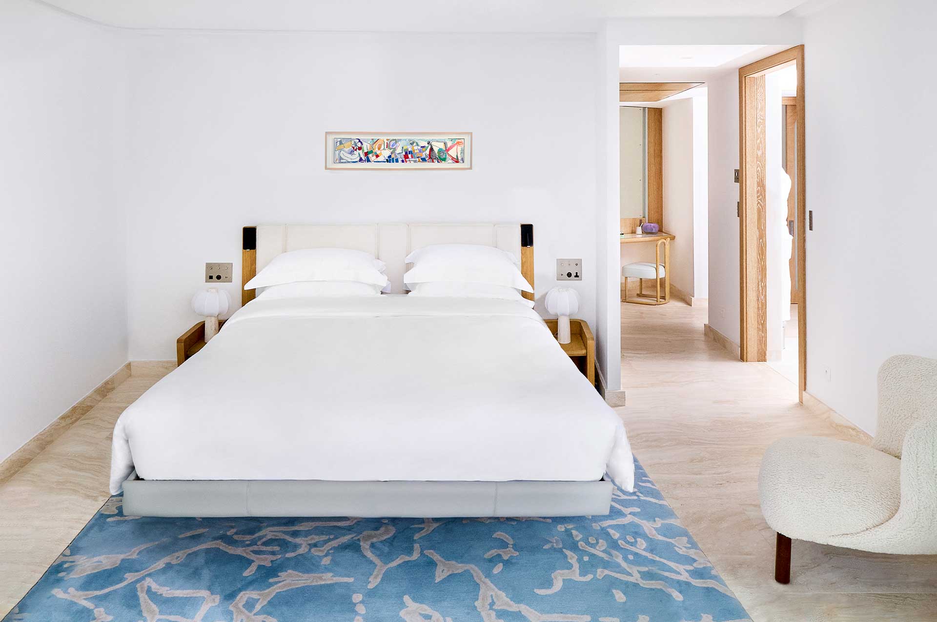 The Sea View Suite features a large white bed in the centre with some colourful artwork hanging above the bed. The open door on the right leads to the brightly lit, wooden dressing area.