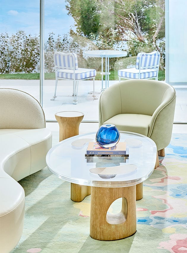 A white leather sofa and armchair with a glass table near an open terrace overlooking a garden and trees.