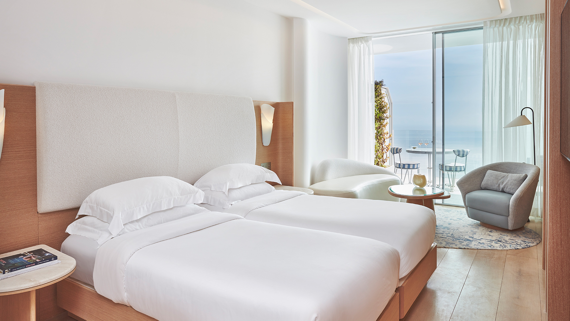 Two singe beds side by side in the Corniche Room with a lounge area and terrace in the background.