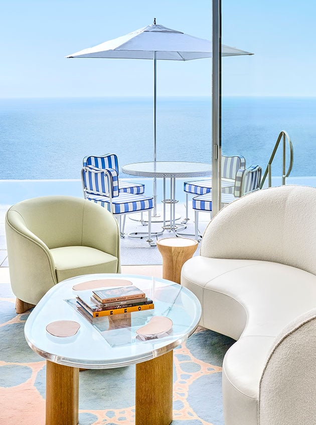 A white leather sofa and armchair with a glass table near an open terrace overlooking a garden and trees.