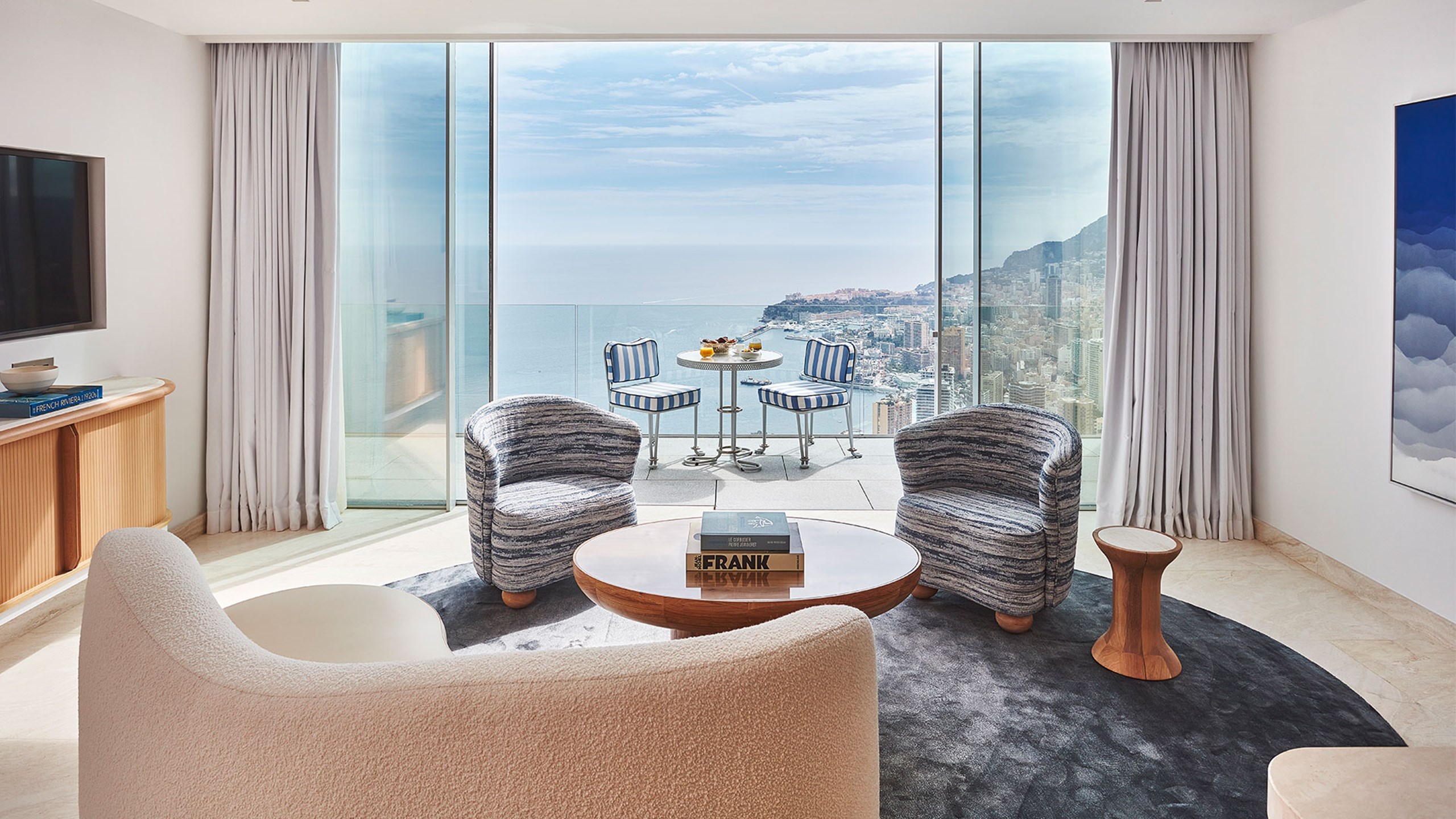 Le salon de la Grande Suite Duplex Azur avec une table basse et deux fauteuils autour, et la terrasse en fond avec une table et deux chaises autour, et la vue sur la Côte d'Azur.