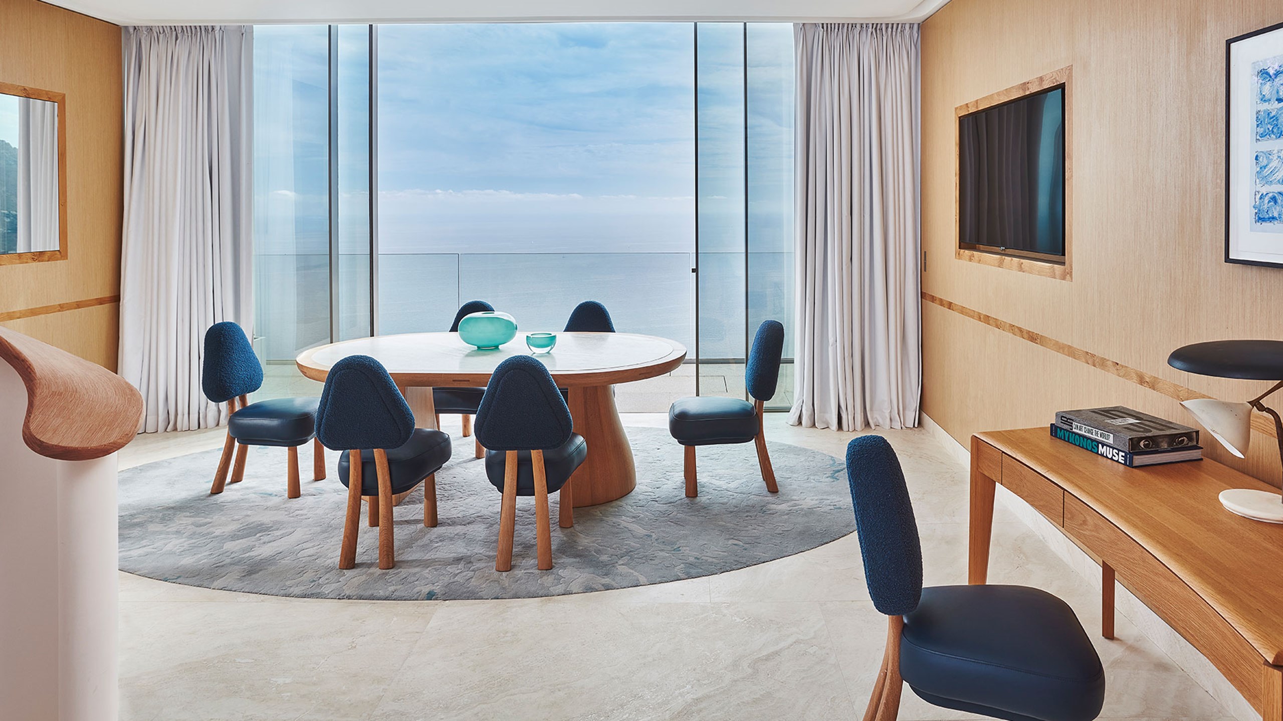 La salle à manger de la Grande Suite Duplex Azur avec une table à manger et des chaises bleu foncé autour, un bureau sur la droite et la terrasse avec la vue sur la mer en fond.