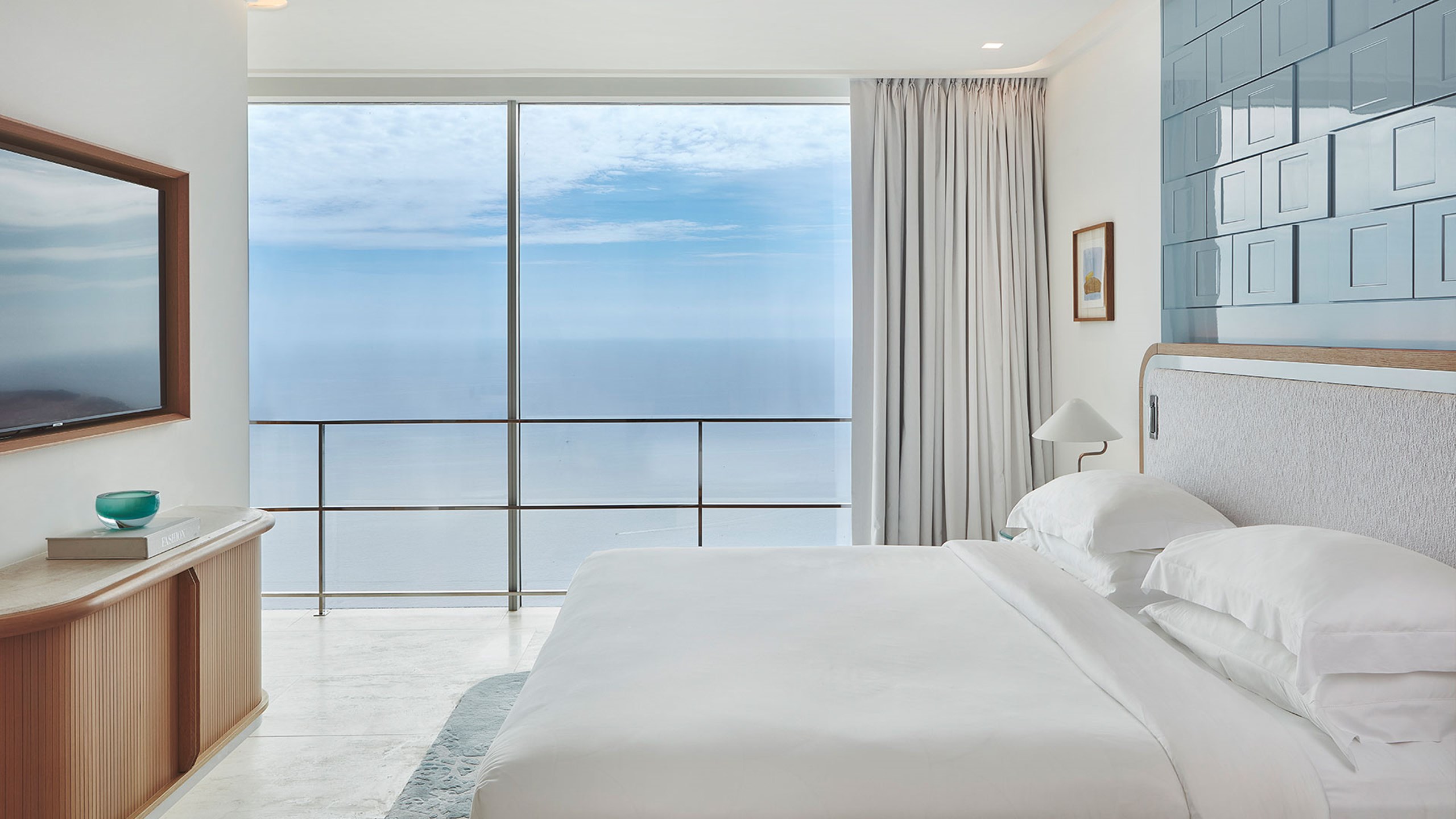 King size bed with white bedding set against a blue tiled wall with patio doors open looking out to sweeping sea view