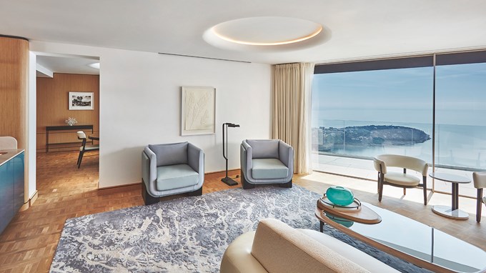 Le salon de la Grande Riviera Suite avec une table basse, deux fauteuils bleu ciel, et la terrasse sur la droite avec la mer en arrière-plan.