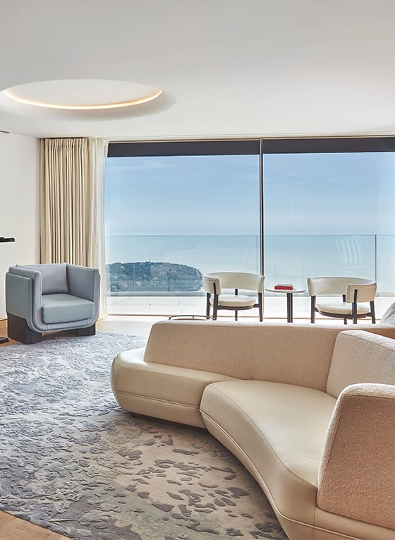 Grand Riviera Suite living room with a long white sofa, two light-blue armchairs, a table with chairs and a terrace with a view on the sea.