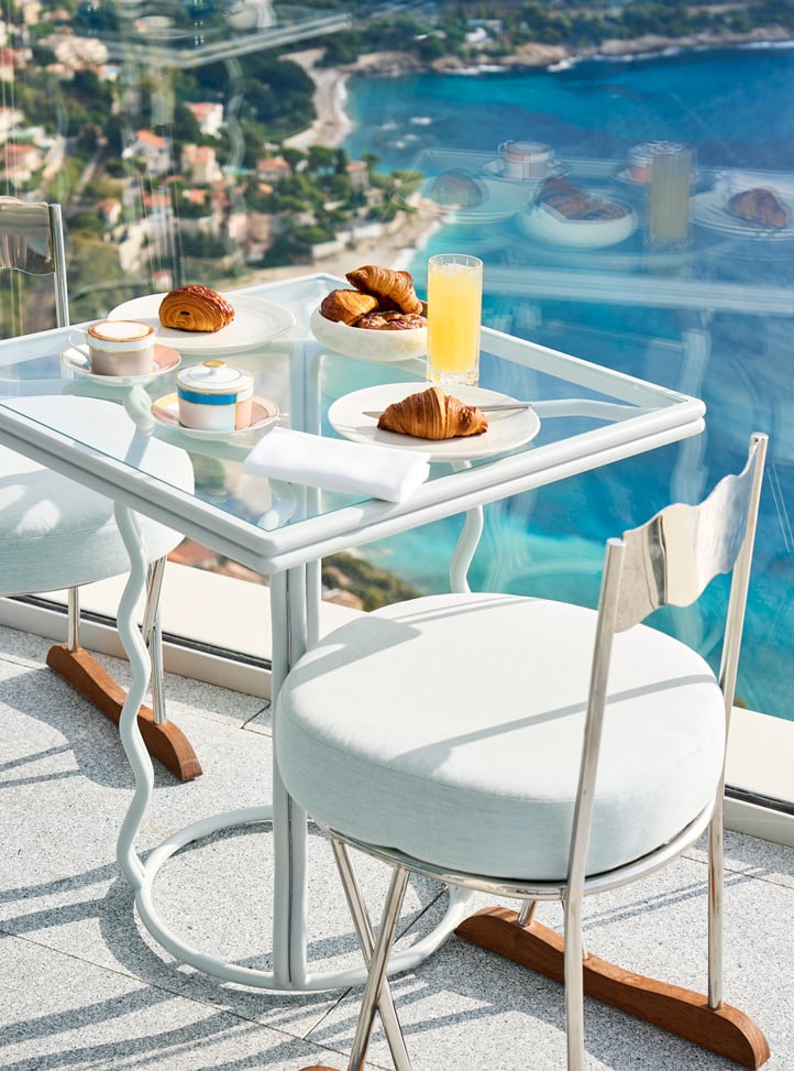 Table on the terrace with croissants and two white chairs with a view of the sea.