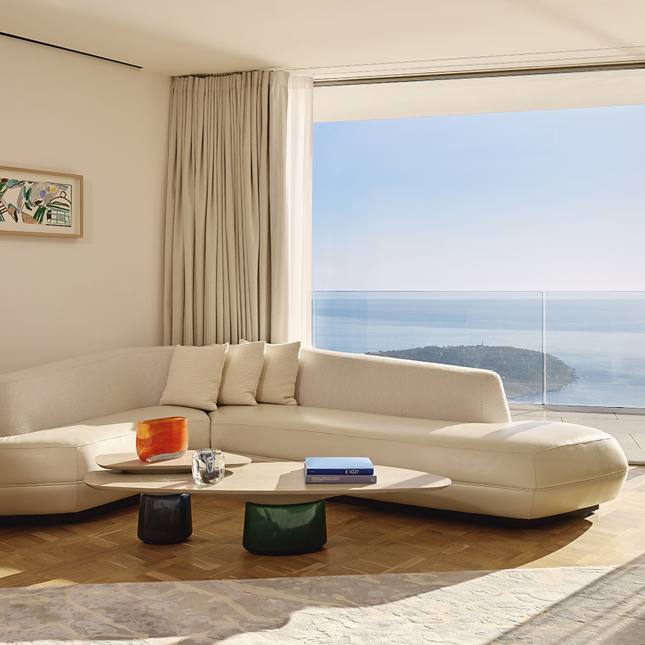 Elegant living area with a curved cream sofa and neutral cushions, two sculptural coffee tables with colourful bases, and a large window offering sweeping sea views, framed by floor-length curtains and parquet flooring.