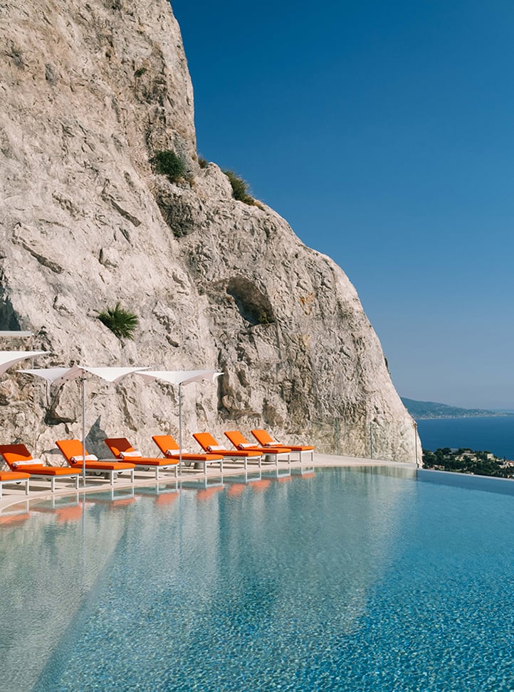 Infinity pool built against a rocky cliffside, with bright orange sun loungers and white umbrellas lined up along the edge, overlooking a sweeping coastal view under a clear blue sky.