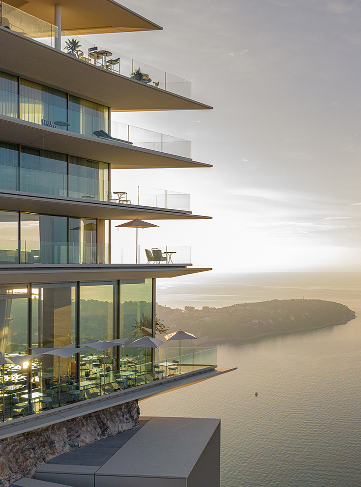 View of the side of The Maybourne Riviera hotel with Riviera Restaurant on the ground floor, rooms and suites above, and the Riviera landscape with the sea in the background.