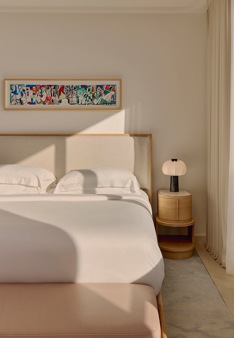 Bright bedroom with a double bed dressed in white linens, a light wood headboard, and a rounded wooden bedside table holding a sculptural lamp, with soft natural light and a colourful artwork above the bed.