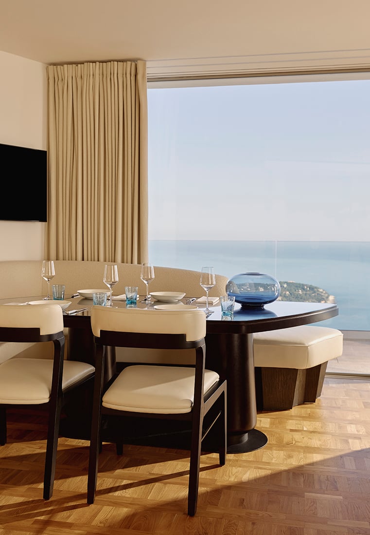 Elegant dining area with a dark wood table set for six, featuring cream-upholstered chairs and a curved banquette, positioned beside a floor-to-ceiling window offering expansive sea views and natural light.
