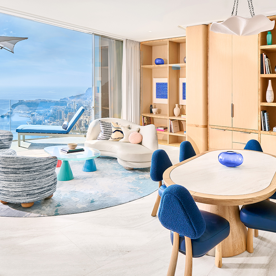 Living room area with a curved white sofa and a coffee table in the centre, a dining table with blue chairs on the right-hand side and a terrace with a blue lounger in the back.