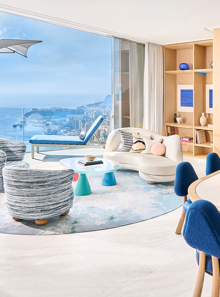 Living room area with a curved white sofa and a coffee table in the centre, a dining table with blue chairs on the right-hand side and a terrace with a blue lounger in the back.