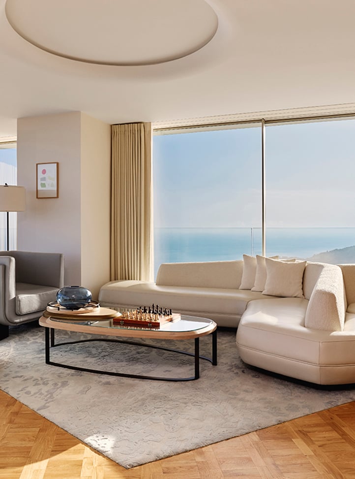 Sophisticated living room with a curved cream sofa, matching armchair, and a modern oval coffee table set on a patterned rug, featuring a chess set and glass bowl, with floor-to-ceiling windows offering panoramic sea and coastal views.