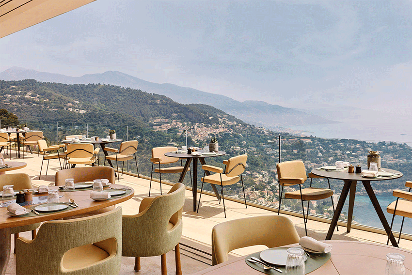 Terrasse d’un restaurant en hauteur avec chaises modernes beige et vert et tables rondes dressées, offrant une vue panoramique sur la côte méditerranéenne et les villages perchés.