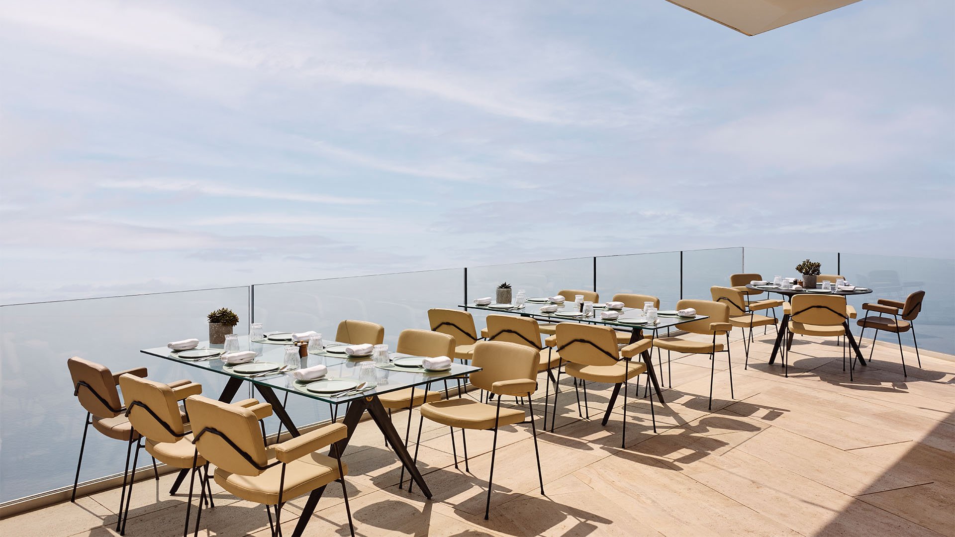 Long tables with beige chairs arranged on a sunlit terrace overlooking the sea, prepared for group dining with neatly set place settings and minimal potted plants.