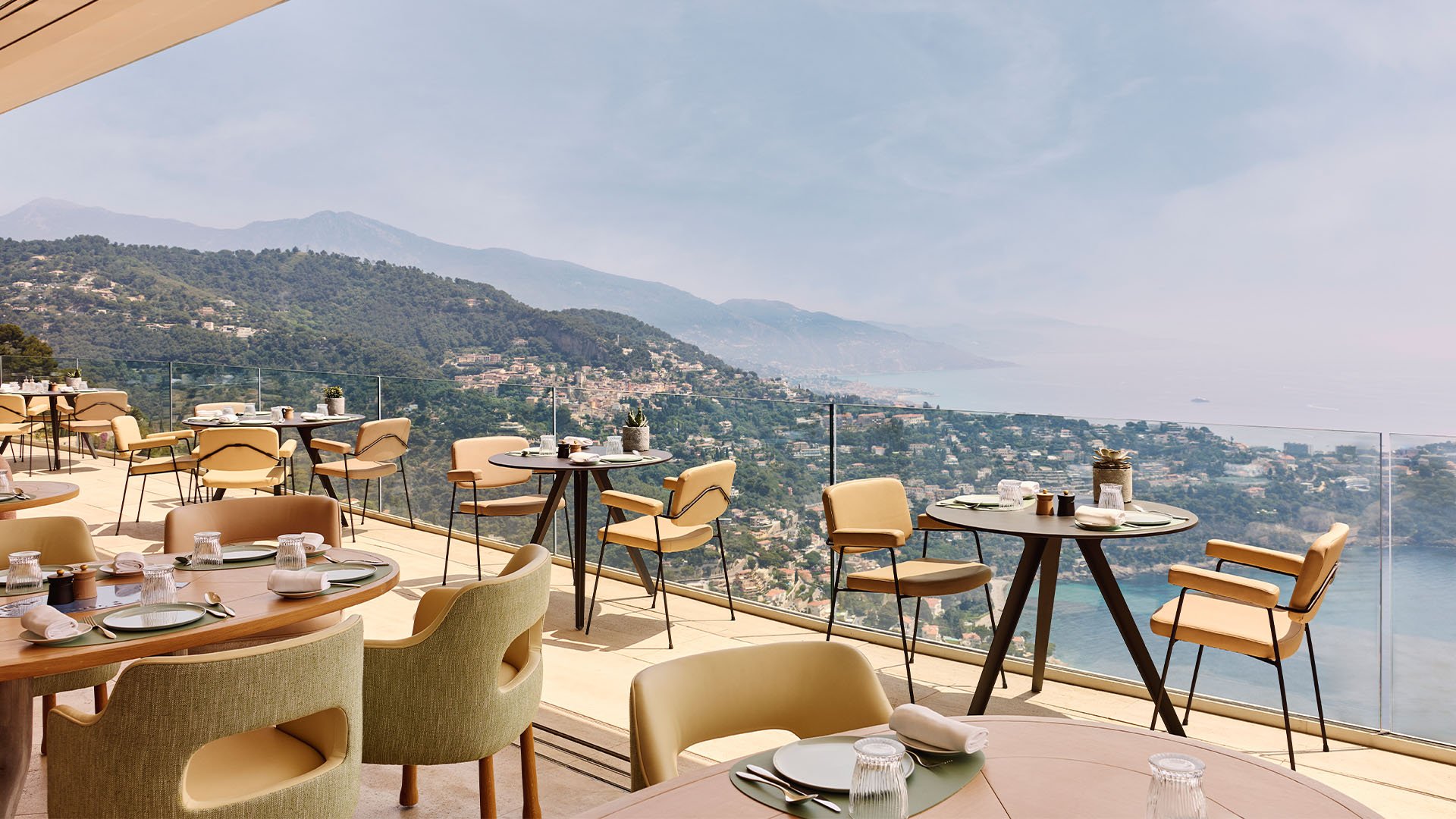 Restaurant terrace with round tables and pale green chairs, offering sweeping views of the coastline, hillside villas, and a hazy blue horizon.
