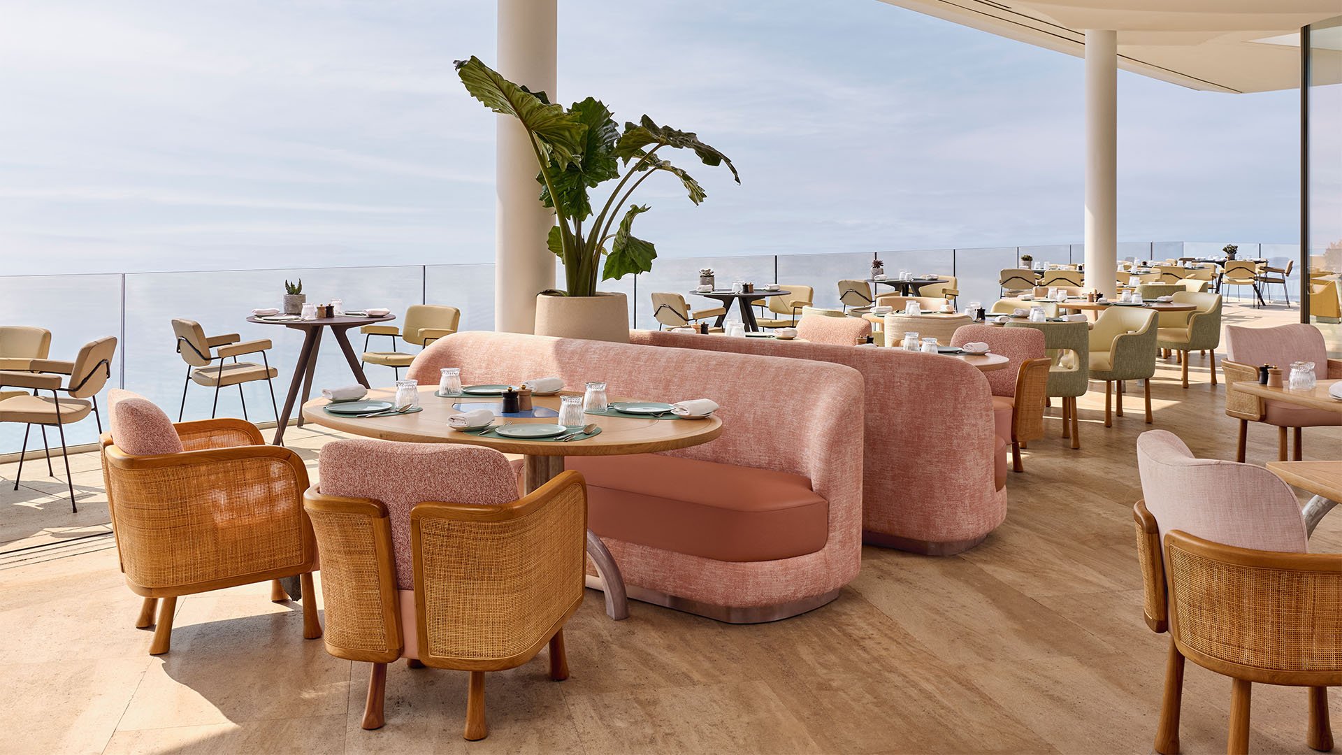 Elegant indoor dining space with soft pink and rattan chairs, ocean views through floor-to-ceiling windows, and pastel-toned decor creating a relaxed, airy atmosphere.