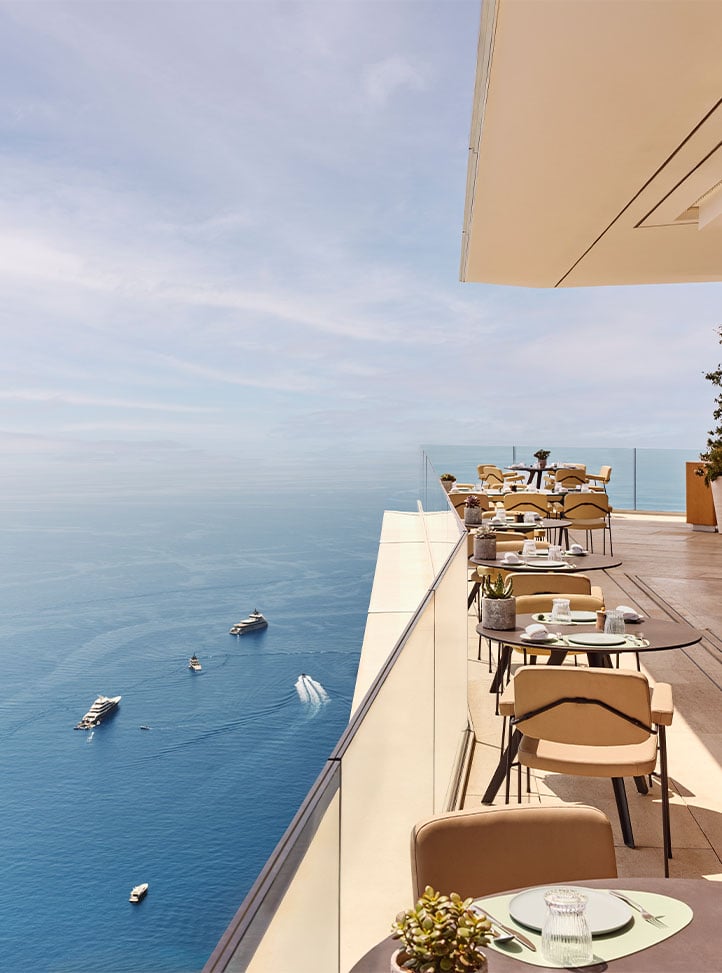 Terrace restaurant overlooking the sea with modern tables and beige chairs, set along a glass balustrade with yachts visible below on the calm blue water.