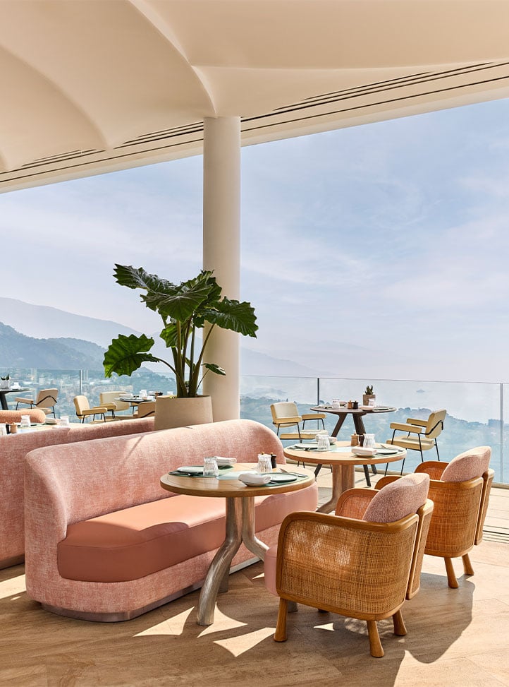 Intimate dining area with peach-toned banquettes and woven armchairs, set beside glass railings with scenic mountain and sea views in the background.
