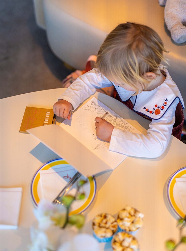 A young child wearing a floral-collared outfit colours in a line drawing with crayons at a small round table indoors.
