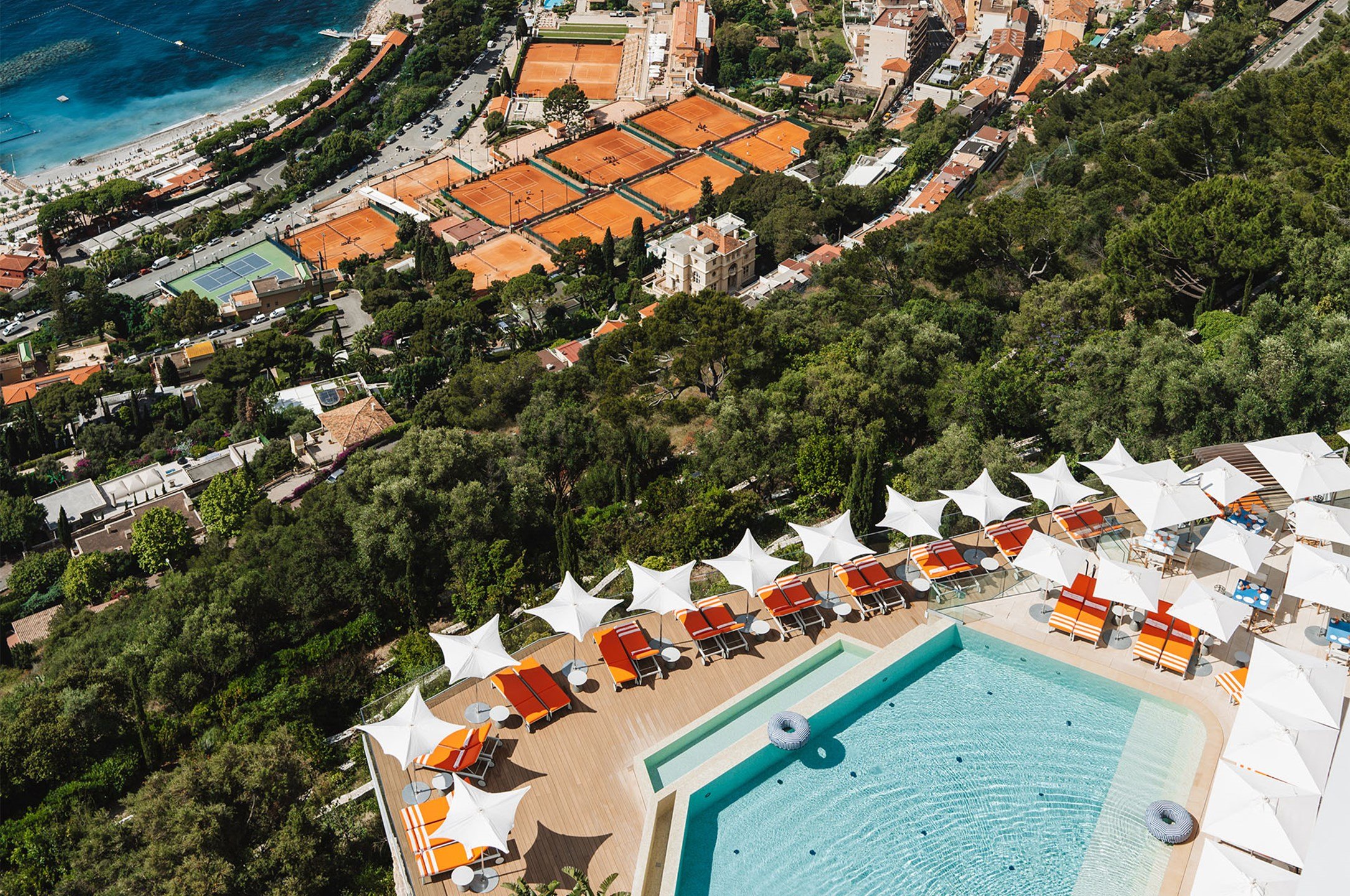 Aerial view of a hillside pool deck with orange loungers and white parasols, overlooking tennis courts and the coastline below.