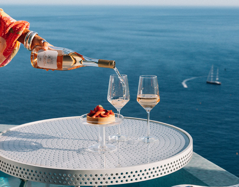 Woman's hand pouring two glasses of rose, with a strawberry tart placed beside the glasses. The sea and boats in the background.