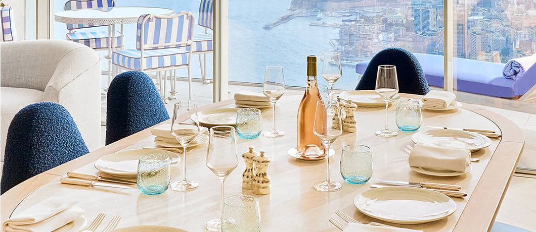 table laid with plates, cutlery, napkins, water glasses, wine glasses and a bottle of rose in the centre of the table. The view overlooks Monaco
